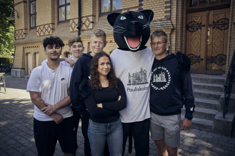 Ett gäng glada elever och Paulimaskot i en gruppbild framför skolan. I bakgrunden syns skolans husfasad. 