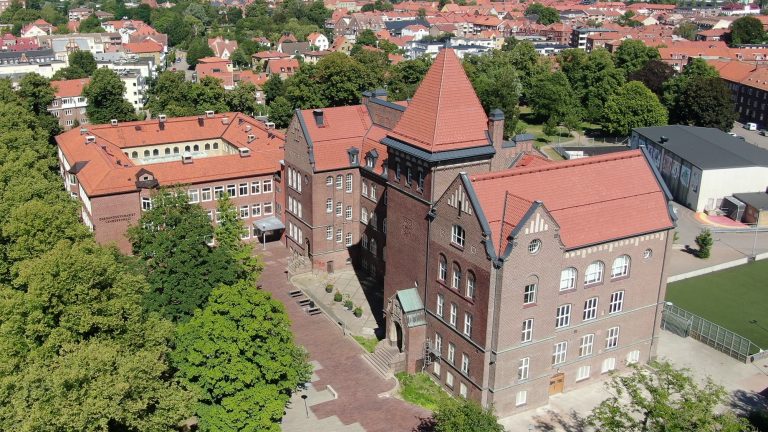 Stor röd tegelbyggnad med träd fotograferad ovanifrån och övrig bebyggelse omkring.