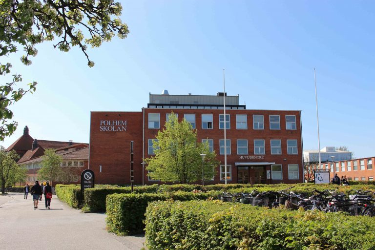 Polhemskolans huvudbyggnad med grönska och cykelställ i förgrunden. Elever syns på trottoaren.