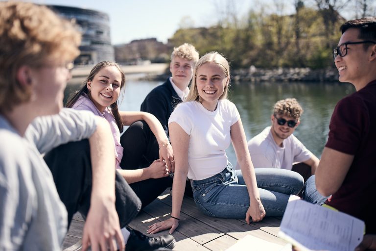 Ett gäng glada elever sitter på kajen vid vattnet utanför skolan och samtalar. I bakgrunden skymtar Hovrätten.