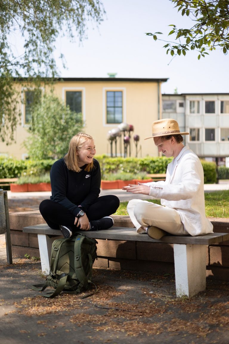 Två glada elever, en flicka och en pojke sitter på en parkbänk och samtalar.