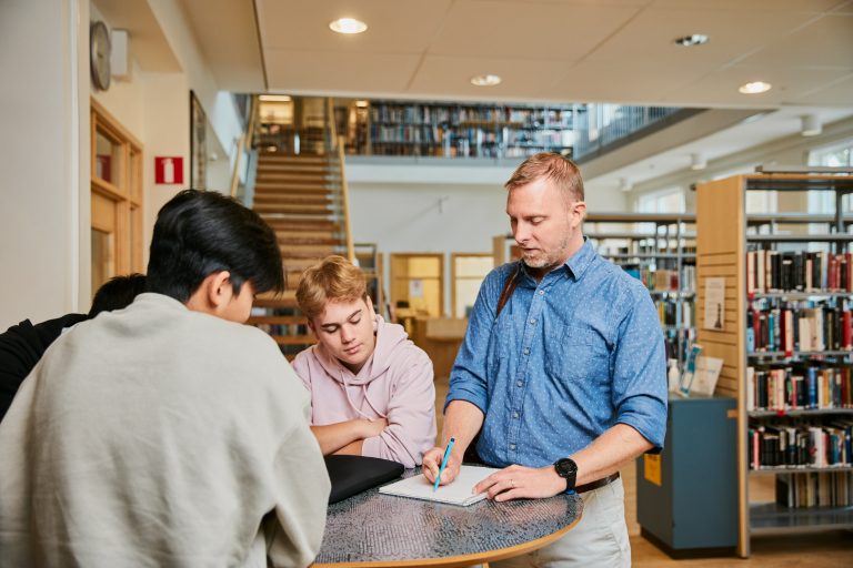 Lärare förklarar en uppgift för två elever som studerar i biblioteket. 