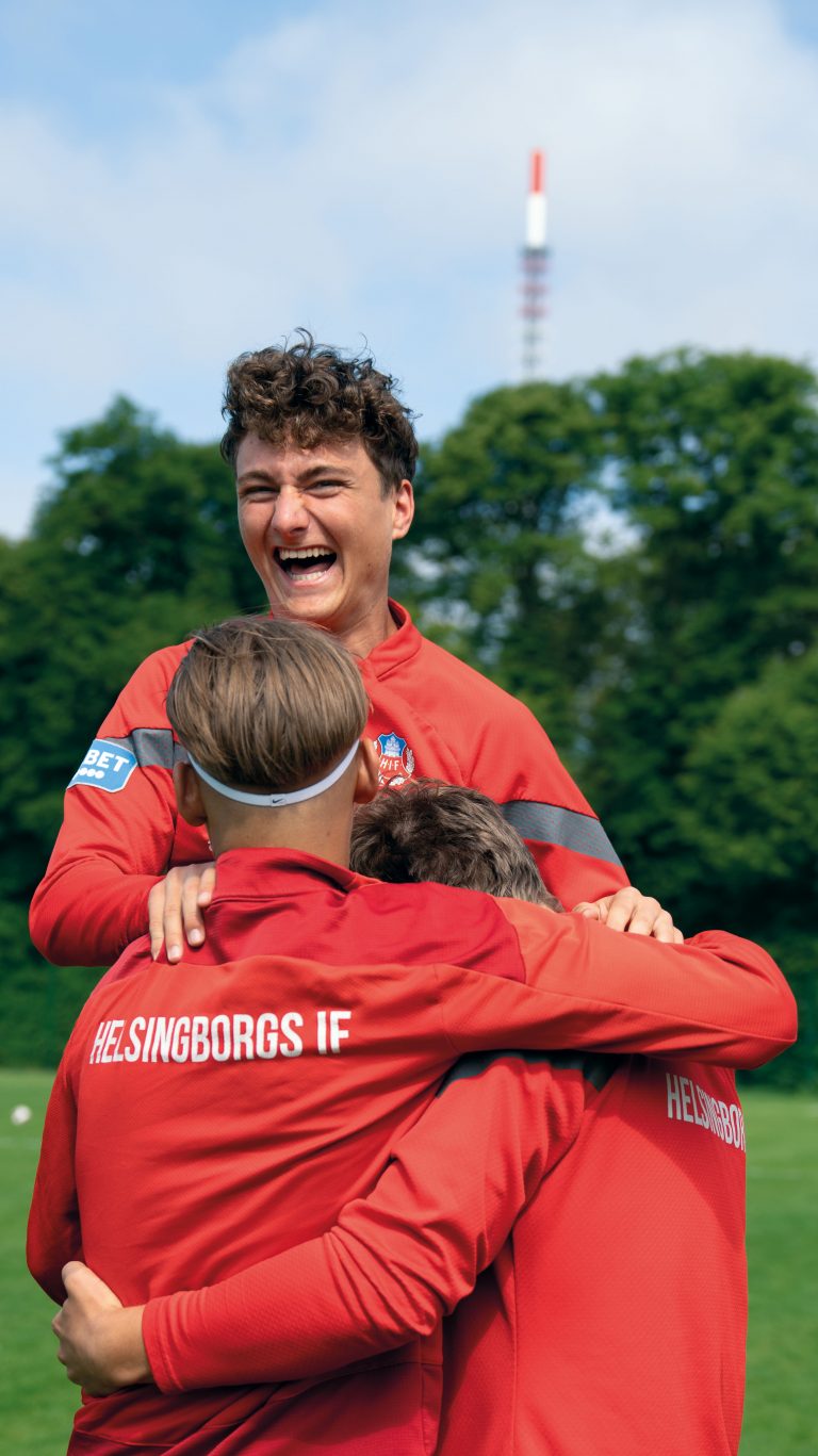 Tre glada fotbollselever håller om varandra ute på en plan med träd i bakgrunden.