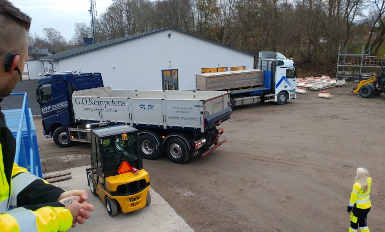 Två lastbilar kör på en grusplan. En gul truck står parkerad. Två personer tittar på.