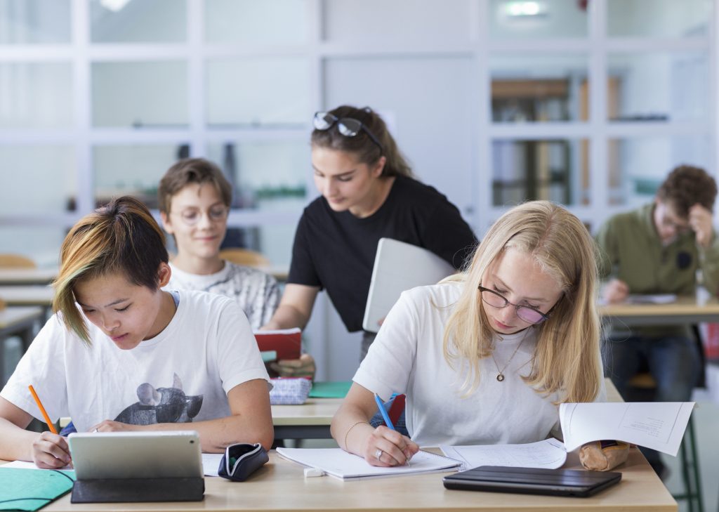 Några elever sitter och arbetar i klassrum. En får hjälp av en lärare.