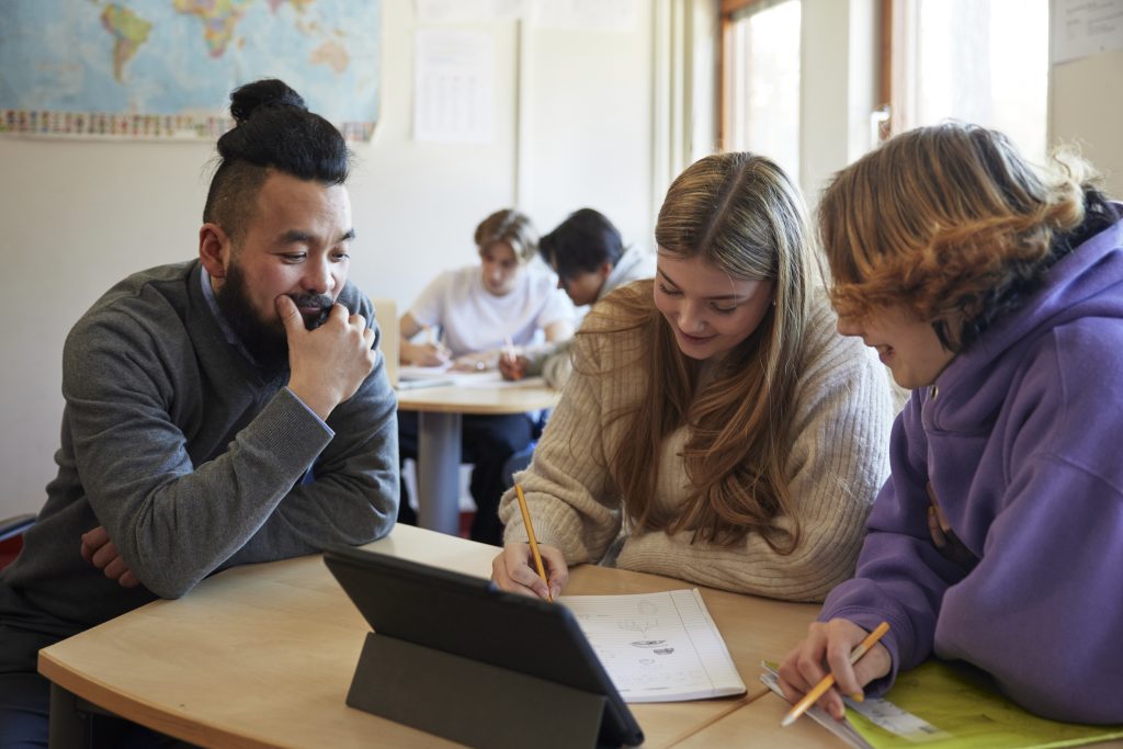 Lärare sitter vid två elever i klassrum. Bakom dem skymtar fler elever.