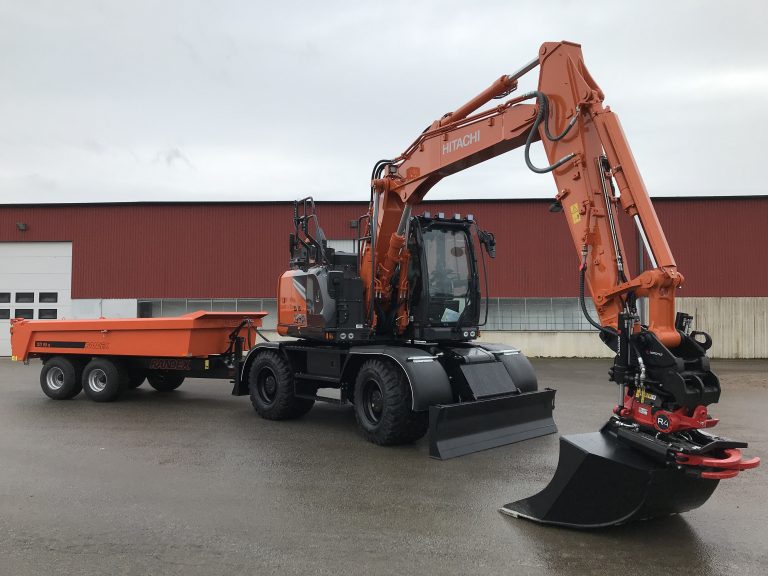 Orange grävmaskin ute på en gårdsplan med en röd byggnad i bakgrunden.