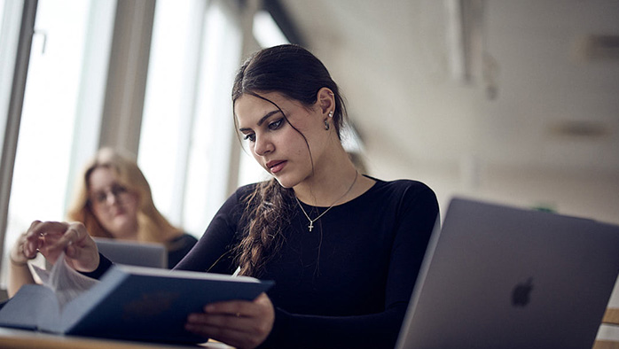 Elev med en laptop och en bok framför sig i ett klassrum med andra elever i bakgrunden.