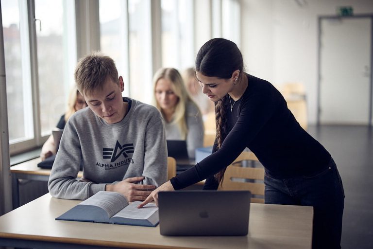 Två elever i ett klassrum med en bok och laptop på bordet framför sig.