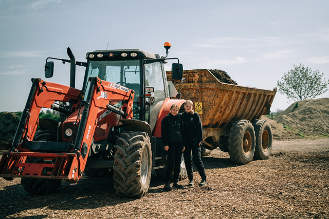 I vårsolen står två leende elever bredvid en röd traktor med släp, redo för praktiskt arbete.