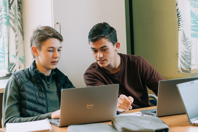 Två elever sitter sida vid sida vid sina laptops i ett ljust klassrum, mitt i ett samtal.