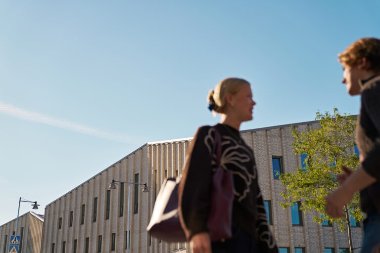 Två personer passerar framför en modern skolbyggnad i ljust tegel. Byggnaden har stora fönster och ligger under klarblå 