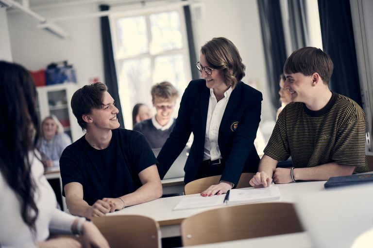 Två elever och en lärare pratar och skrattar tillsammans i ett klassrum där man kan se fler elever i bakgrunden. 