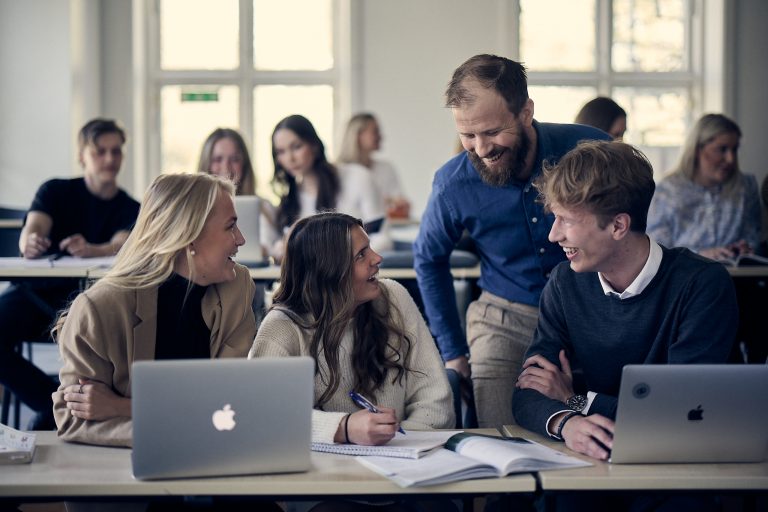 Glada elever arbetar intensivt i ett klassrum. En lärare hjälper tre elever som sitter med dator och skrivböcker. 